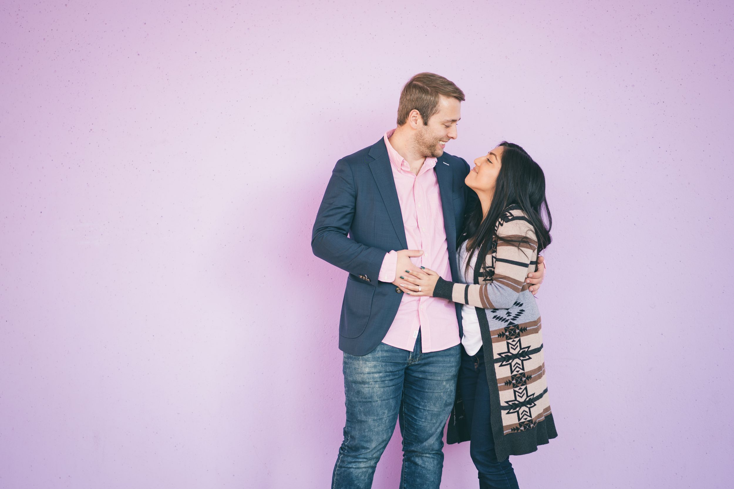 Couple smiles and leans close together against a soft purple background during engagement session.