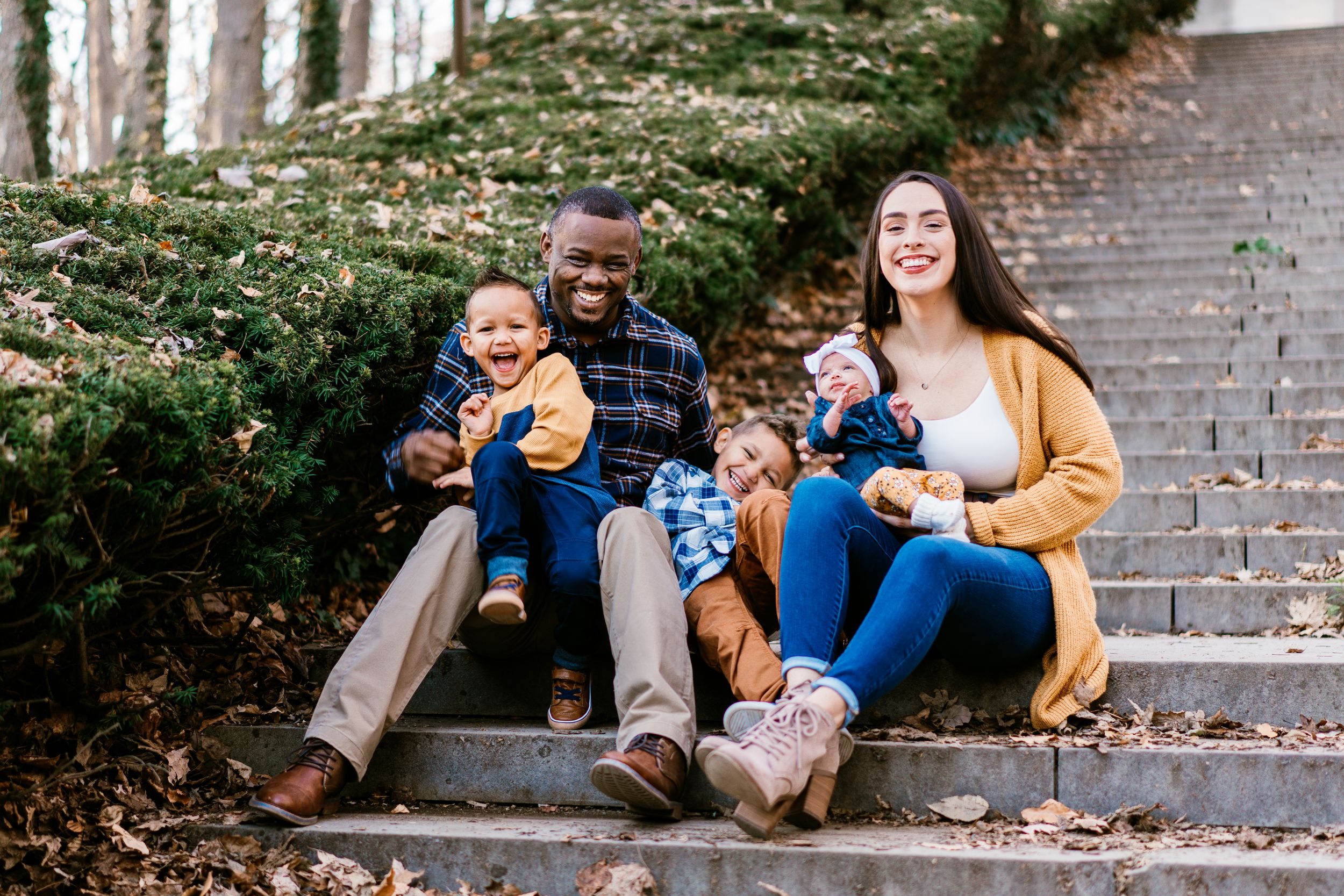 Happy interracial family with two young children sitting on outdoor steps surrounded by autumn leaves.
