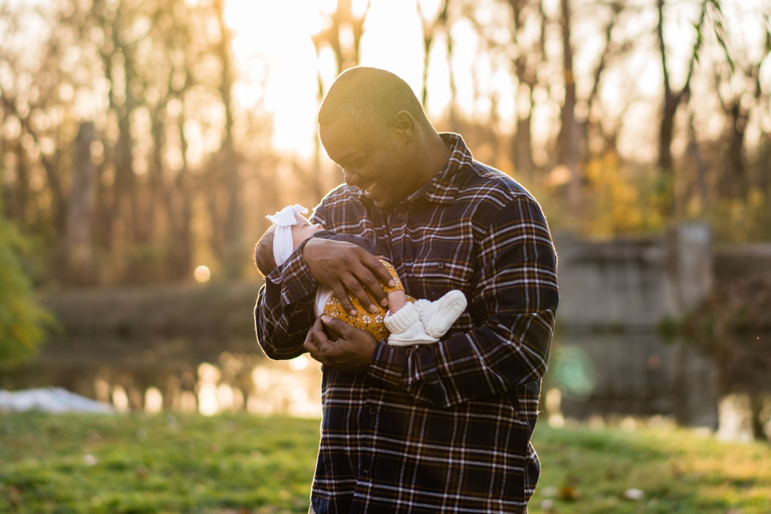 Father tenderly holds newborn baby outdoors at golden hour near a peaceful pond.