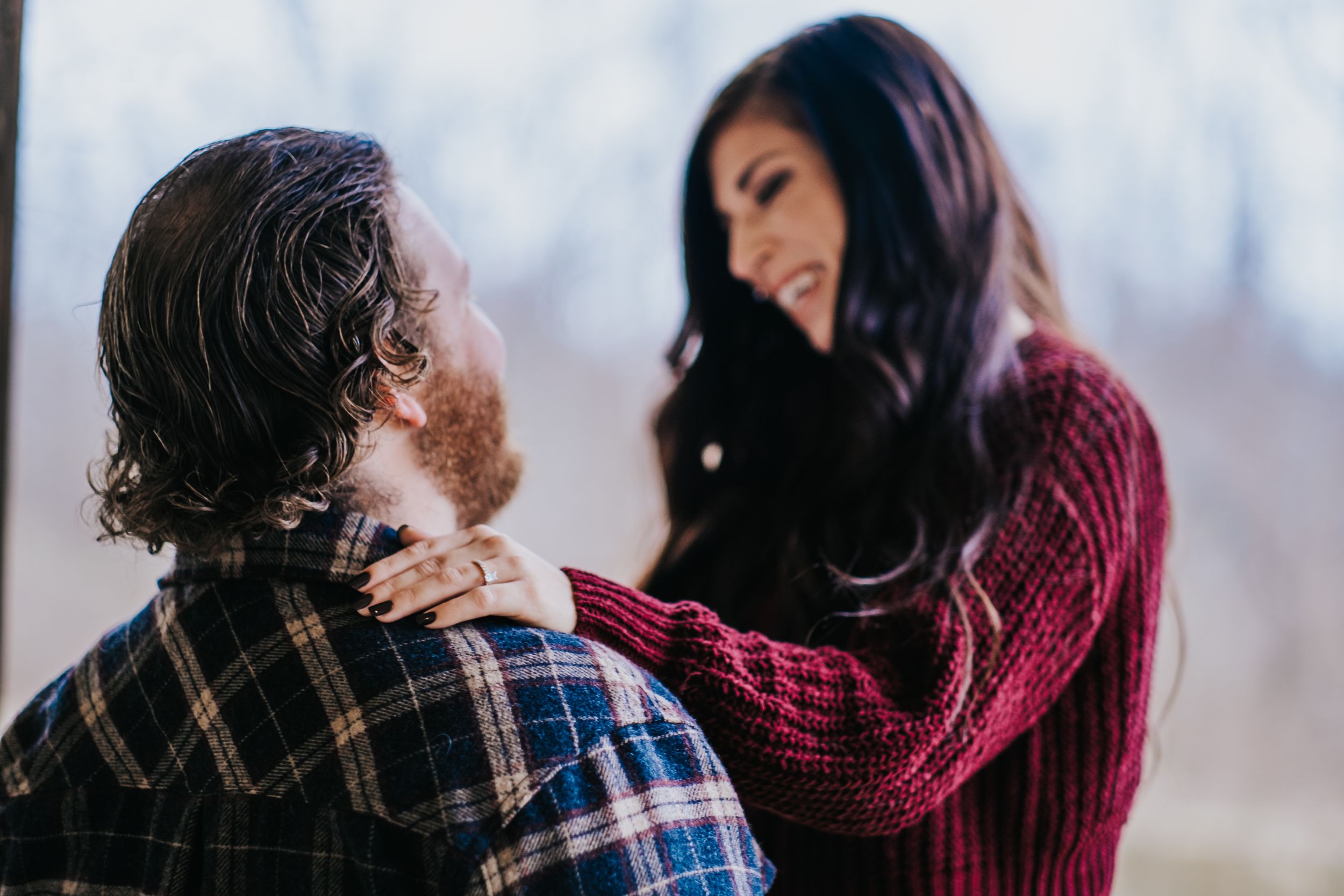 Woman in burgundy sweater smiles warmly at bearded man during intimate winter engagement photo session.