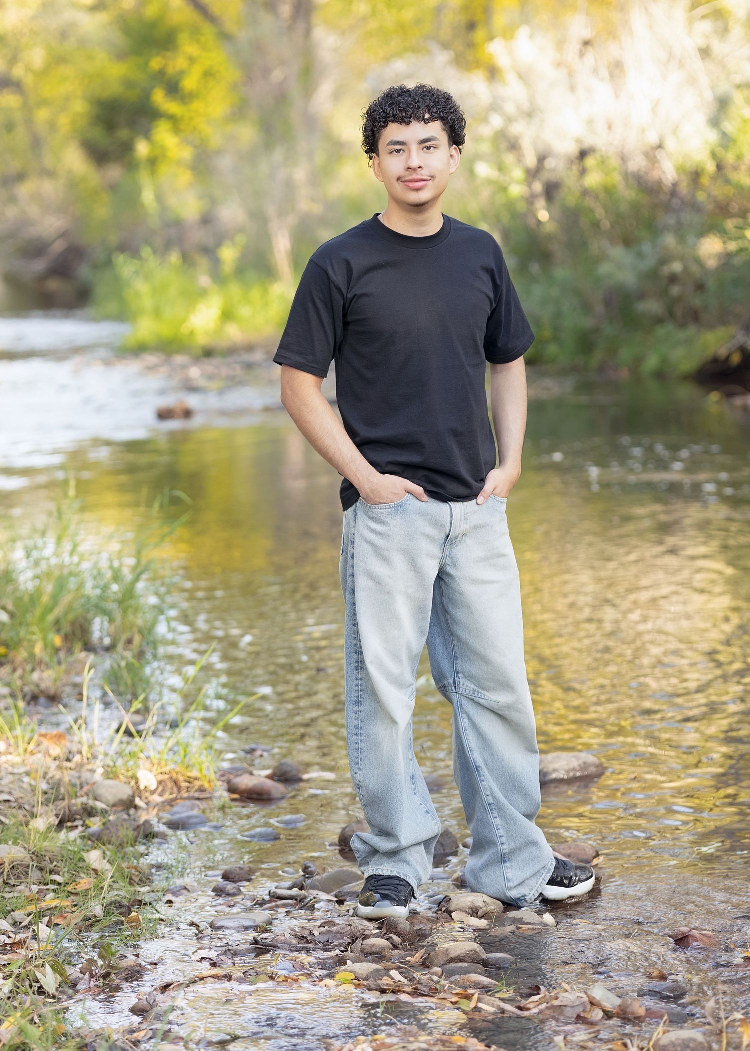 Young man in black t-shirt and light jeans standing on rocks by a stream in autumn.