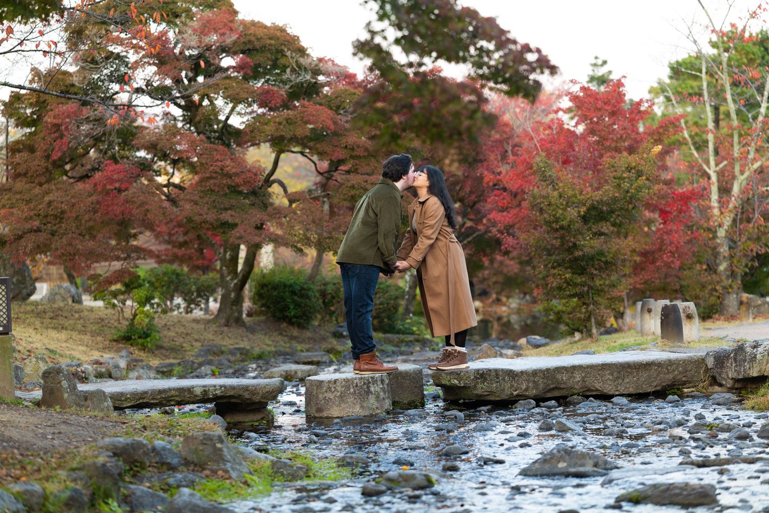 Couple kissing on stepping stones over a stream in a Japanese garden with vibrant autumn foliage.