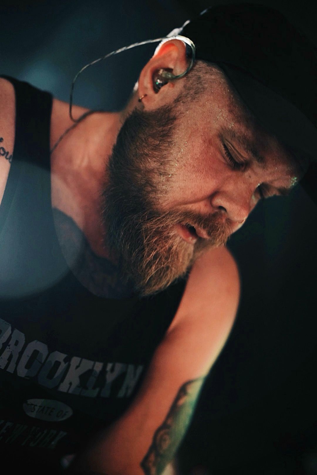 Bearded man wearing a Brooklyn tank top and cap with in-ear monitors performs live on stage under dramatic lighting.
