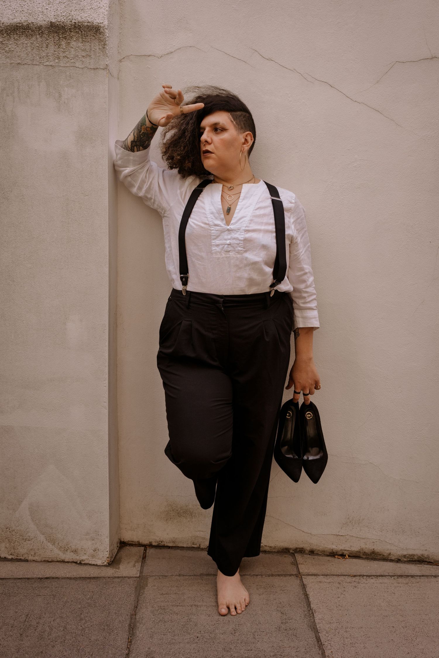 Person in white shirt, suspenders, and black trousers leaning against wall, holding heels barefoot.