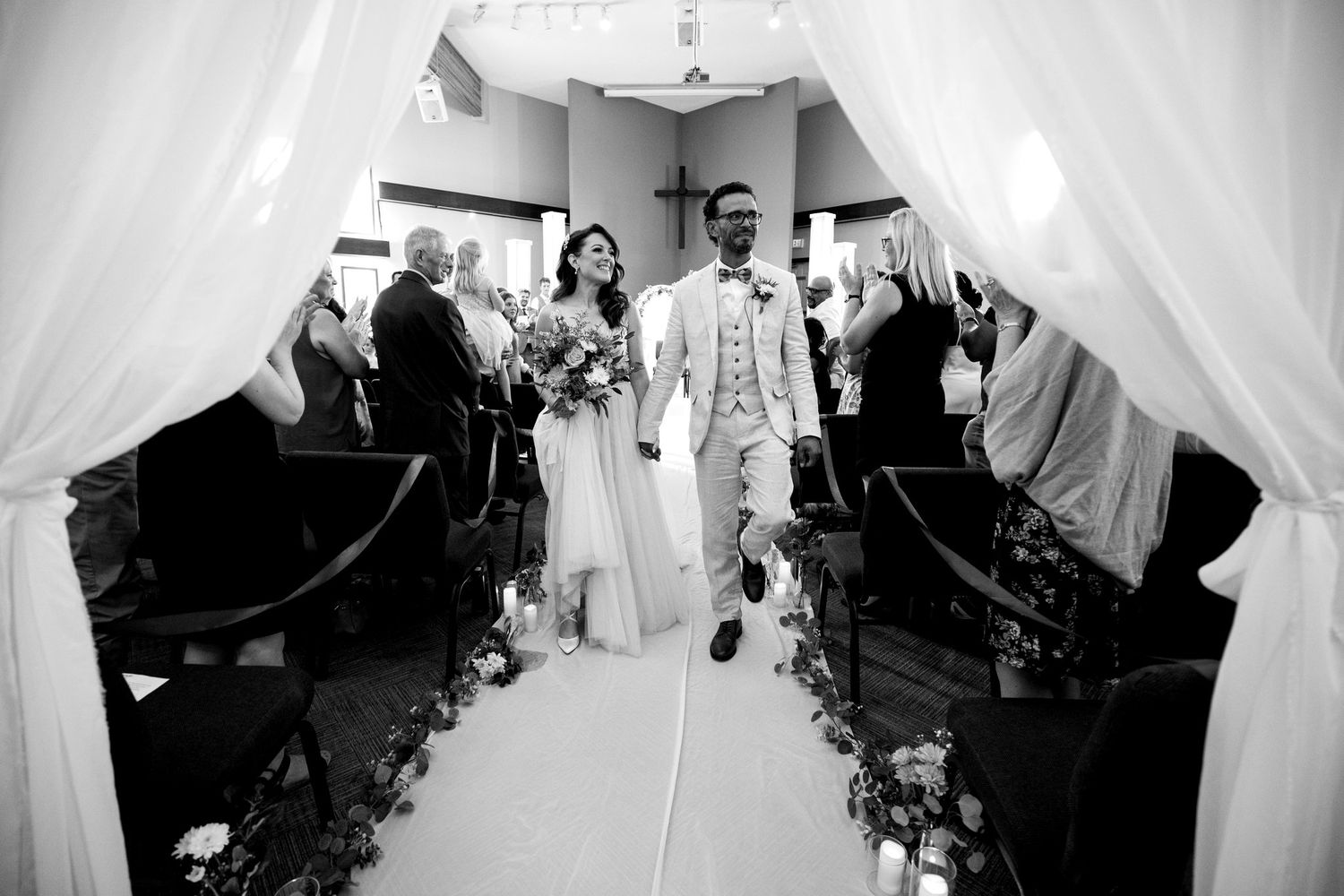 Black and white recessional of the newlyweds walking up the aisle between sheer curtains