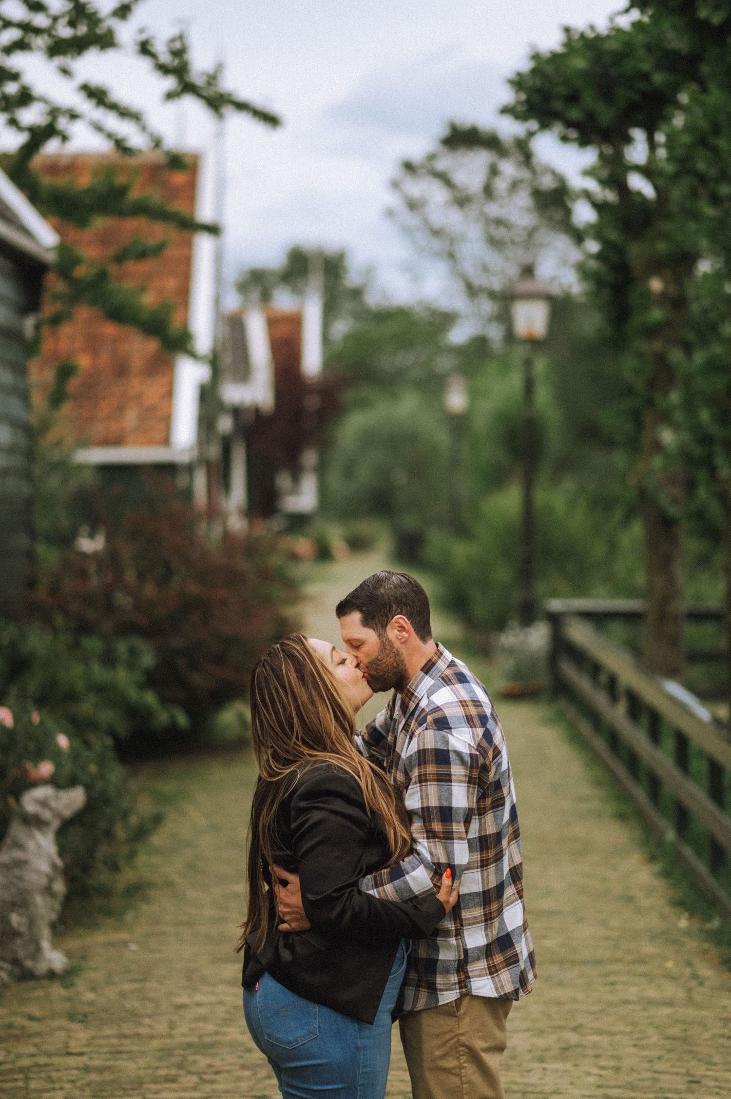 Couple in plaid shirt and black jacket kiss on a brick path lined with greenery, lampposts and a wooden fence in a charming Zaanse Schanse.