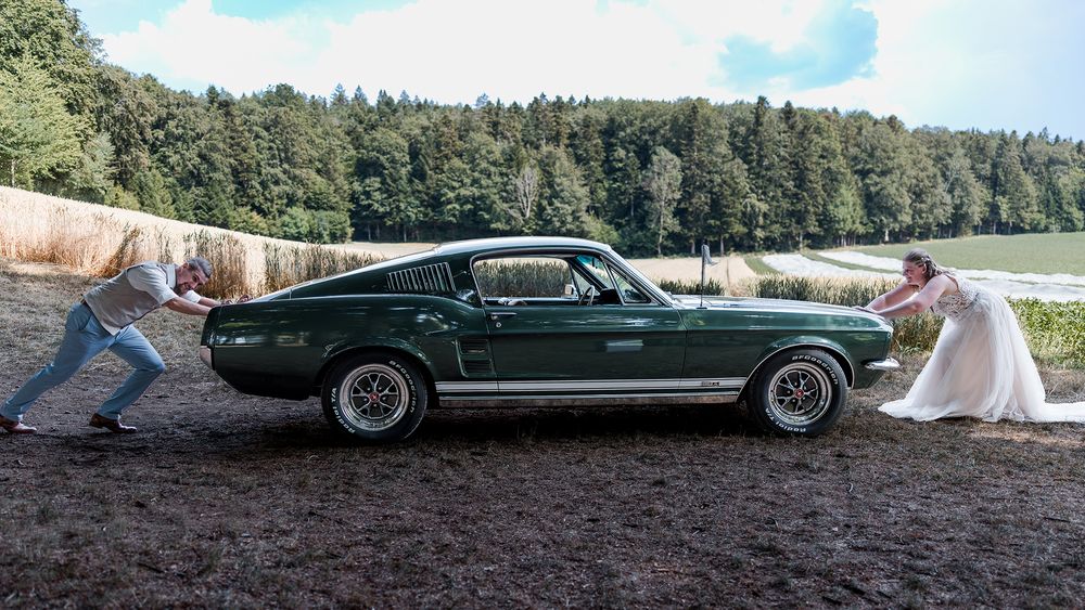 Bride in white gown and man in suit pushing a classic green Ford Mustang fastback on a rural dirt road.
