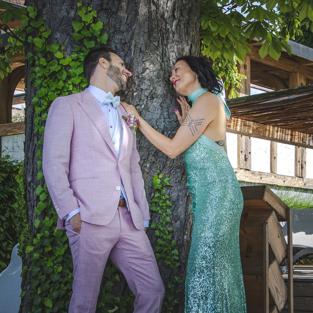 Couple in formal wear posing outdoors, man in pink suit and woman in teal sequin gown adjusting his boutonniere.