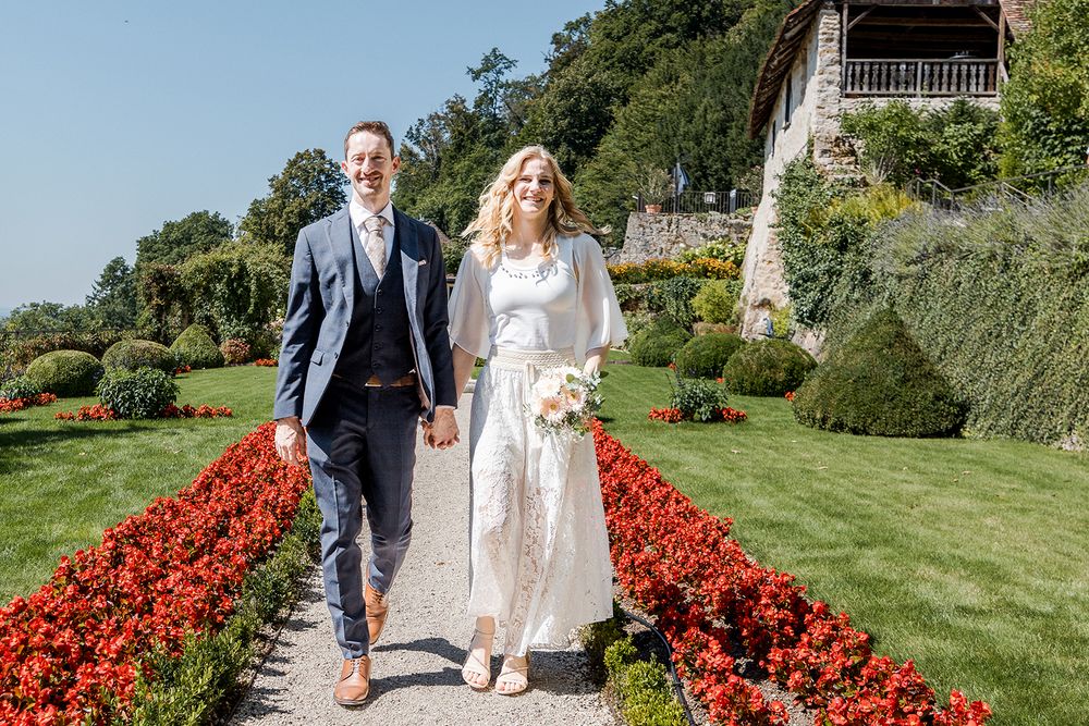 Bride and groom walking hand in hand through a lush garden with red flowers on a sunny wedding day.