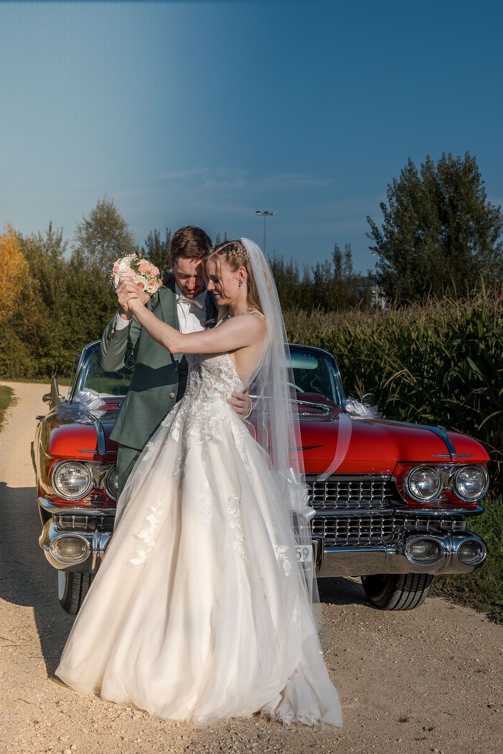 Bride and groom posing together in front of a classic red vintage car on their wedding day.