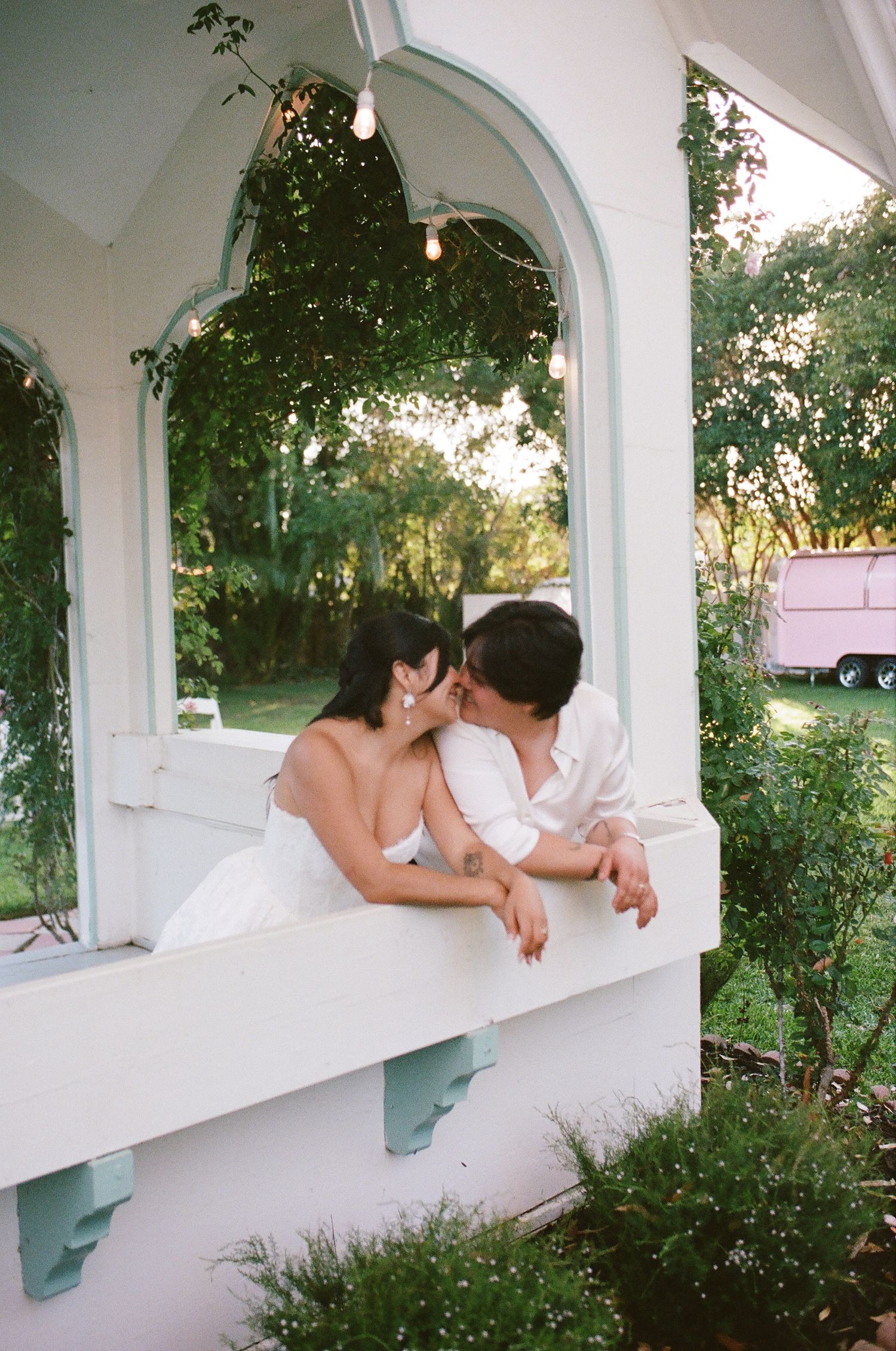 Couple in white outfits sharing a kiss on a white balcony railing surrounded by greenery, with string lights and a pink trailer behind.