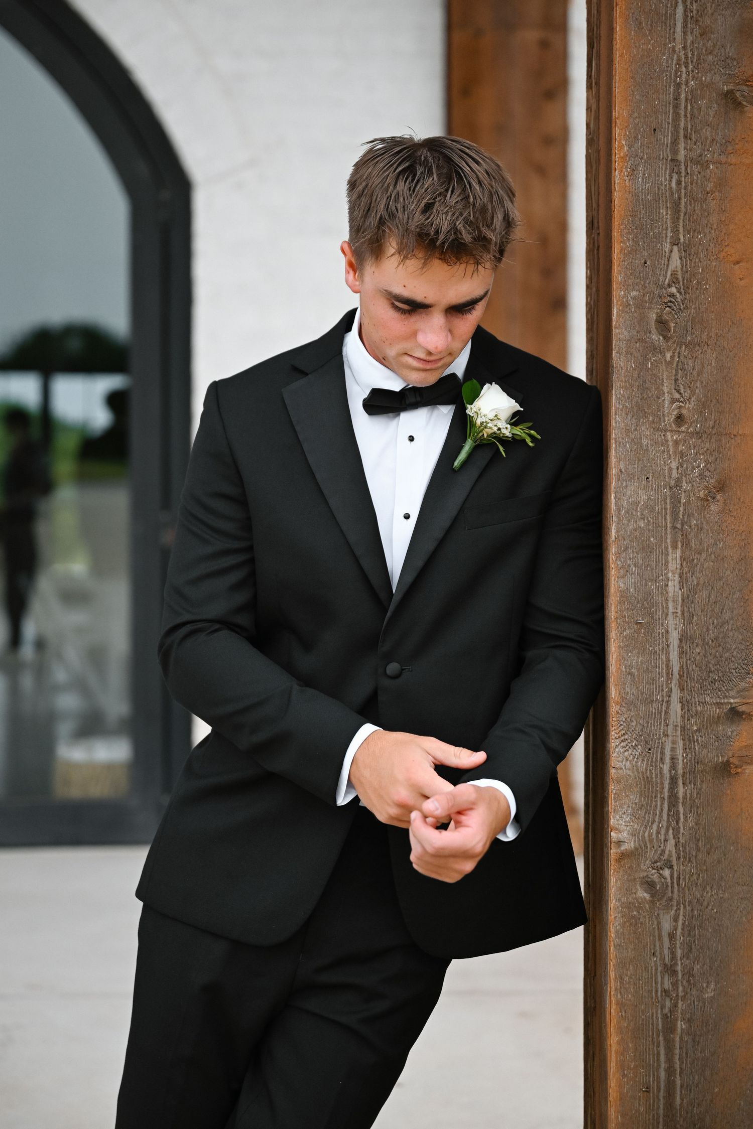 Young groom in black tuxedo with white boutonniere leans against wooden door, adjusting cufflinks before wedding ceremony.