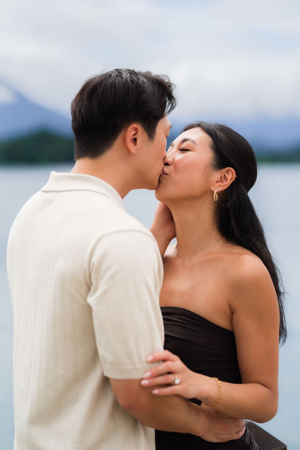 A couple shares a romantic kiss by a serene lake with mountain views, woman in black strapless dress, man in white shirt.