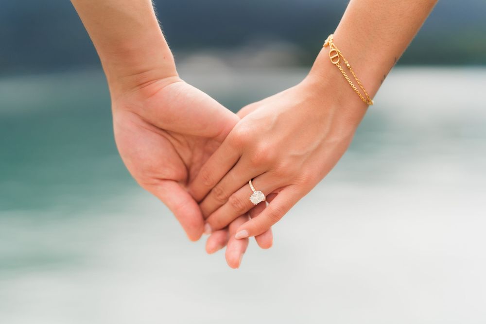 Two hands holding together, one wearing a diamond engagement ring and gold bracelet, with a blurred water background.