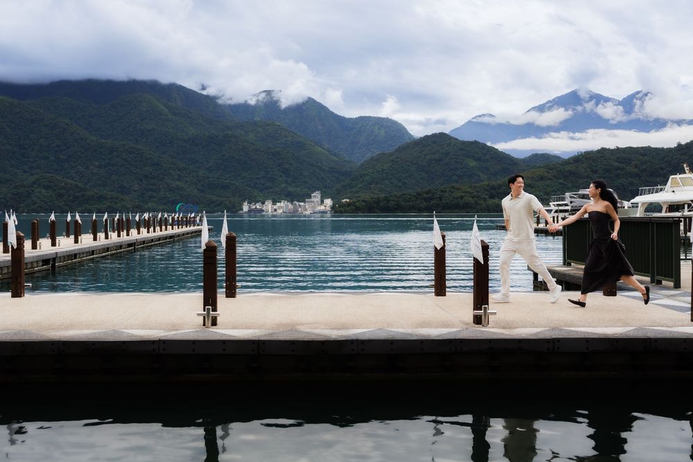 Couple walking on a scenic lakeside dock with lush green mountains and cloudy skies in the background.