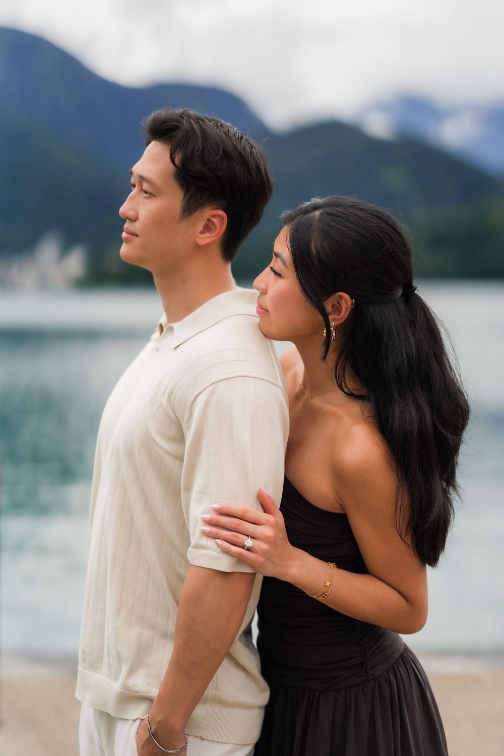 A couple embraces near a lakeside with mountain views, woman in black dress looking up at man in beige shirt.
