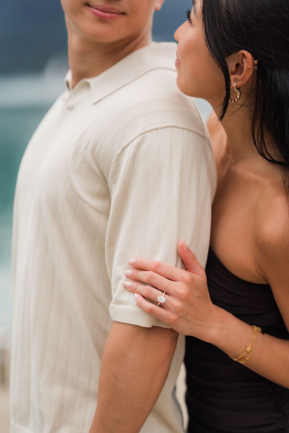 Couple embracing outdoors, woman in black dress with diamond engagement ring and gold bracelet holding man in cream polo shirt.