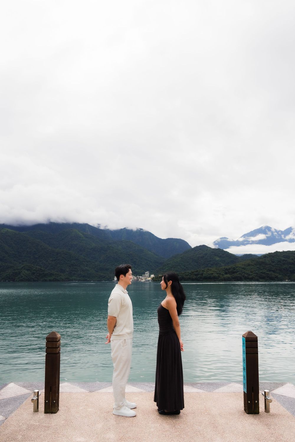 Couple facing each other on a lakeside dock with misty mountains and turquoise water in the background.