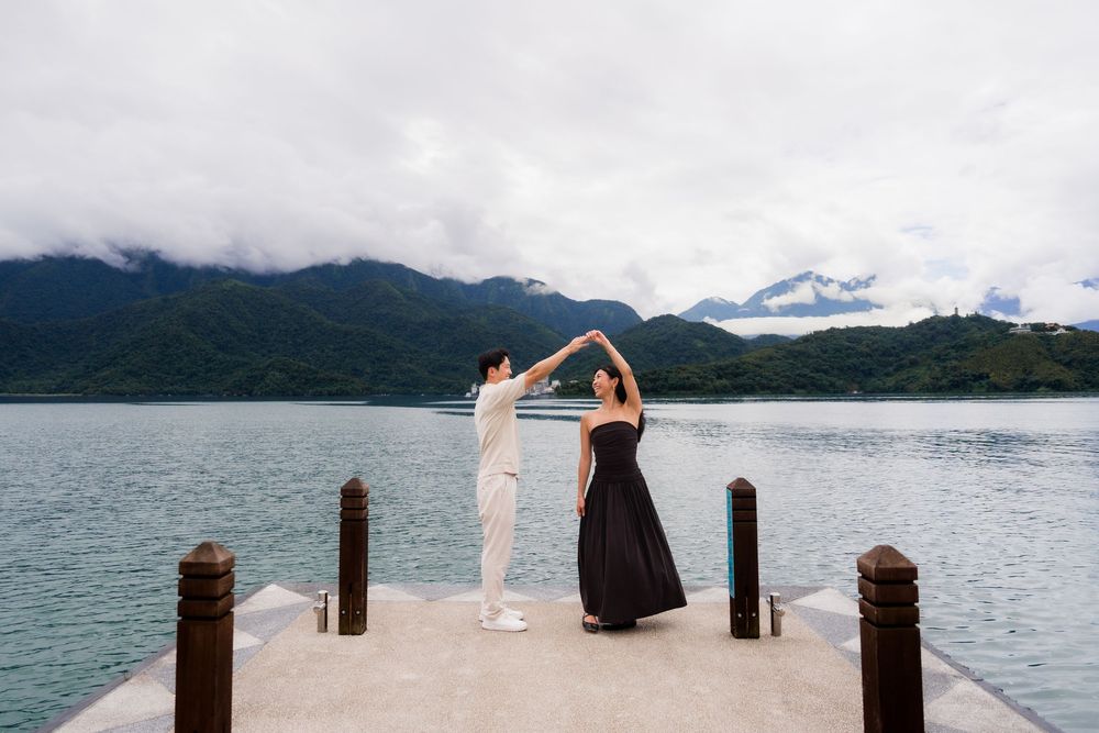 Couple dancing on a lakeside dock with misty mountain backdrop and cloudy skies.