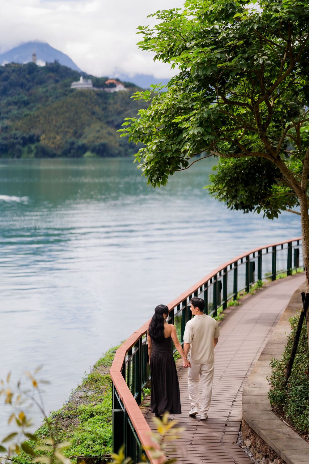 Couple holding hands walking along a scenic lakeside pathway with lush green trees and misty mountains in the background.
