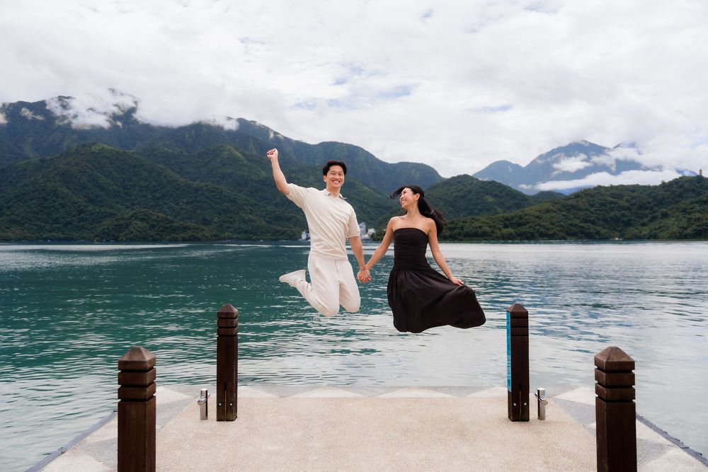 Couple jumping joyfully on a dock with turquoise lake and misty mountains in background.