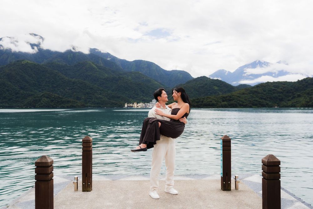 Couple posing on a dock at Sun Moon Lake, man lifting woman with scenic mountains and turquoise water behind them.