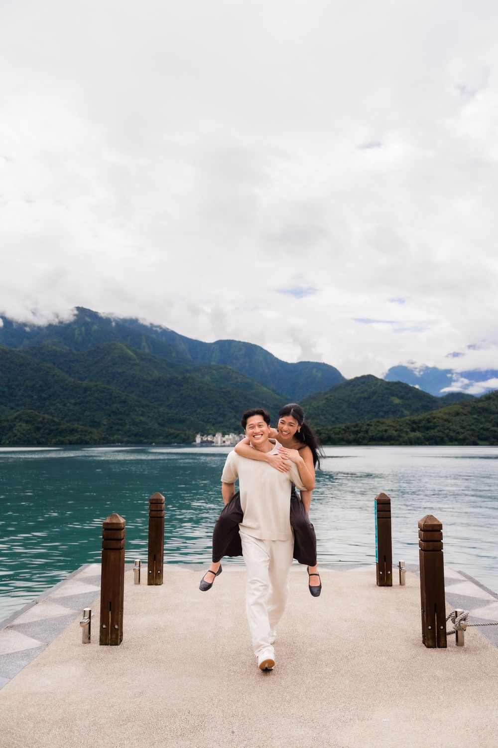 Couple enjoying a piggyback ride on a lakeside pier with lush green mountains and cloudy sky in background.