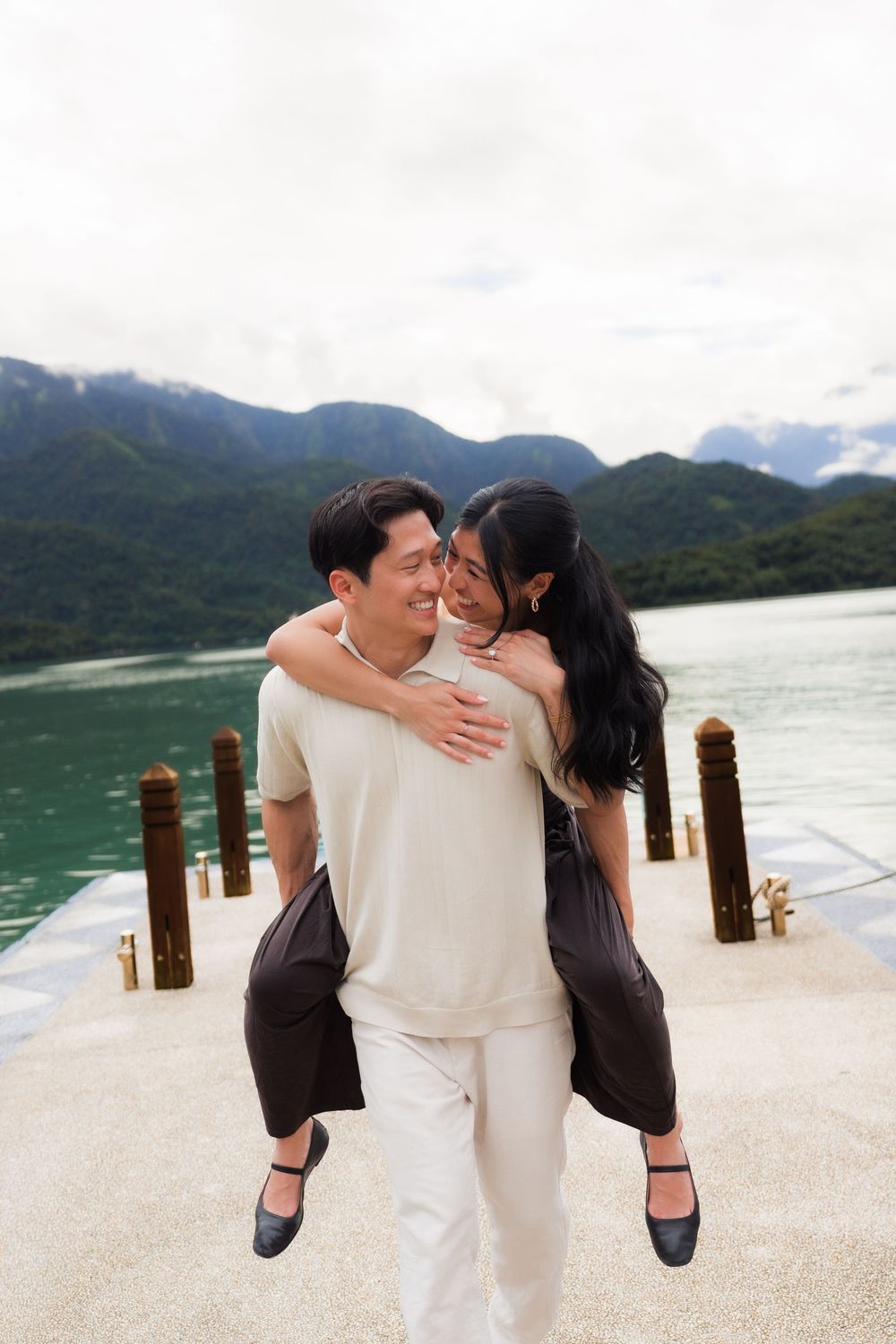 Man giving woman a piggyback ride on a lakeside dock with green mountains behind them.