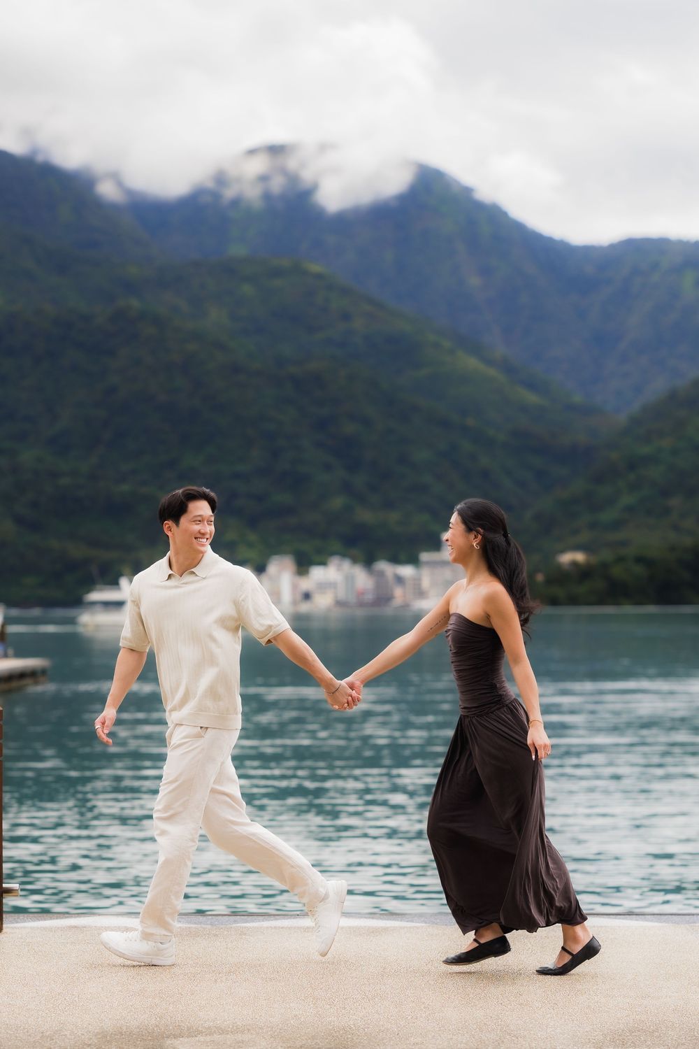 A couple holds hands and walks along a lakeside promenade with lush green mountains in the background.