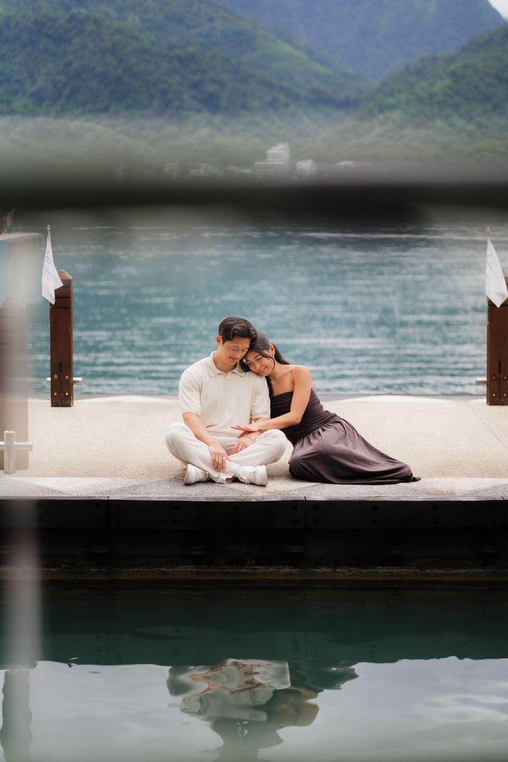 Couple sitting together on a lakeside dock with turquoise water and mountains in the background.