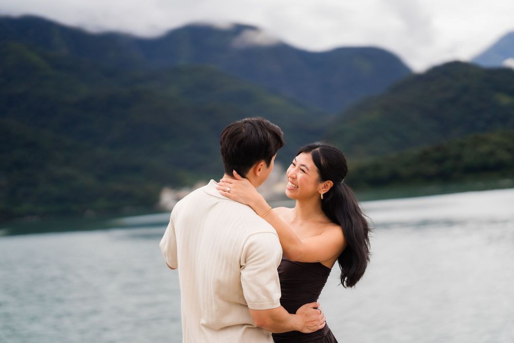 Couple embracing near a serene lake with lush green mountains in the background on a cloudy day.