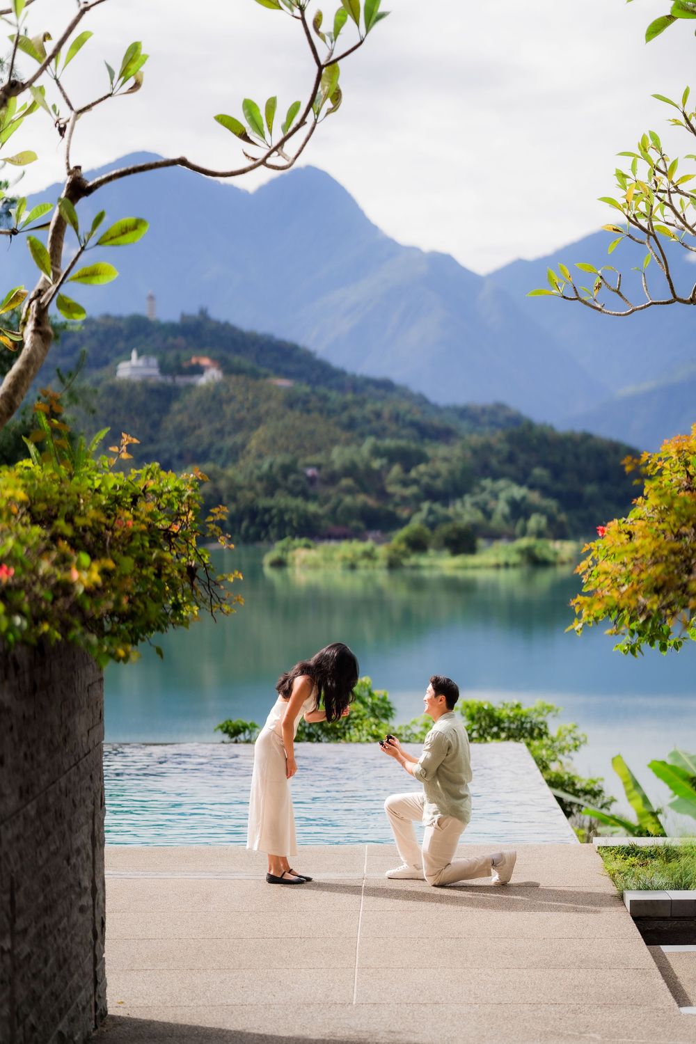 Man proposes on one knee to woman by a serene lake with lush green mountains in the background.