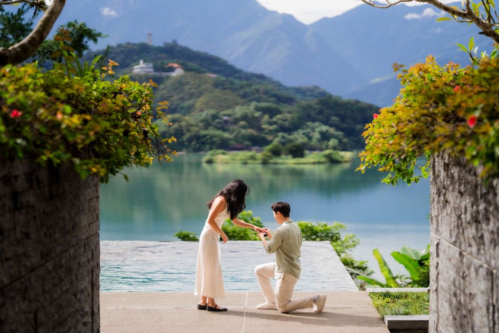 Man proposes on one knee to woman by a serene lake with lush mountains and greenery in the background.