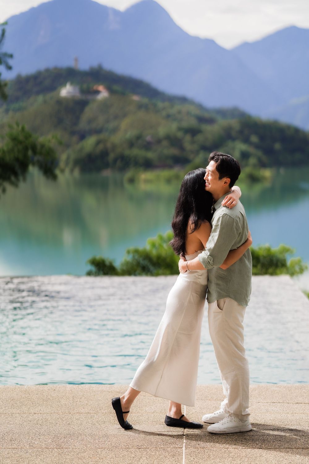 Couple embracing romantically by a turquoise lake with lush green mountains in the background.