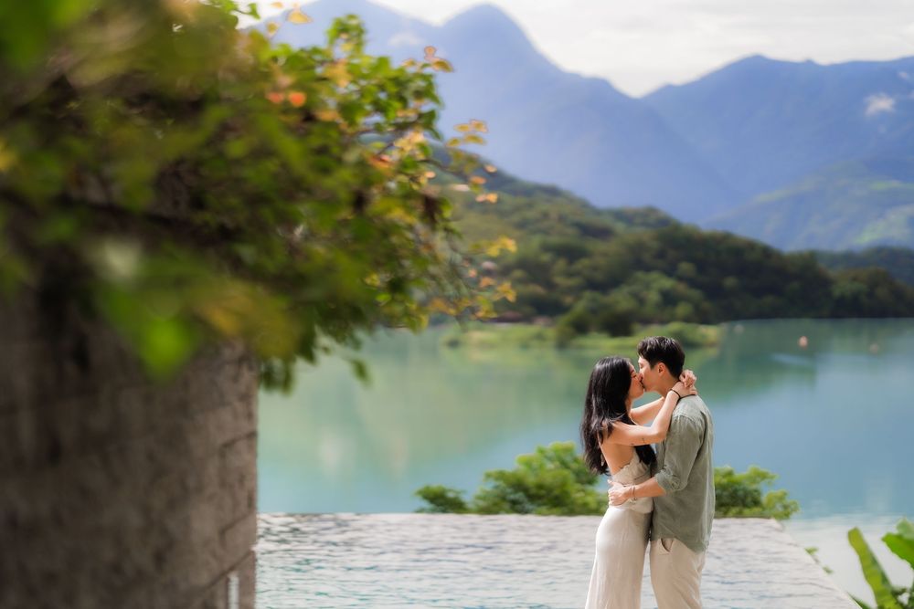 Couple embracing near turquoise lake with lush mountains backdrop in romantic outdoor pre-wedding photo shoot.