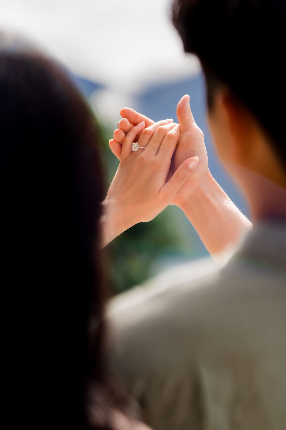 Couple holding hands with an engagement ring visible, shot from behind in a romantic outdoor setting.