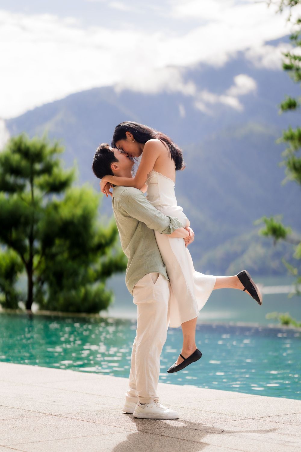 Couple embracing romantically by an infinity pool with mountain lake views on a sunny day.
