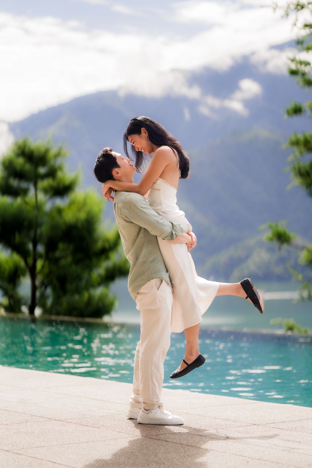 Man lifting woman near infinity pool with mountain backdrop.
