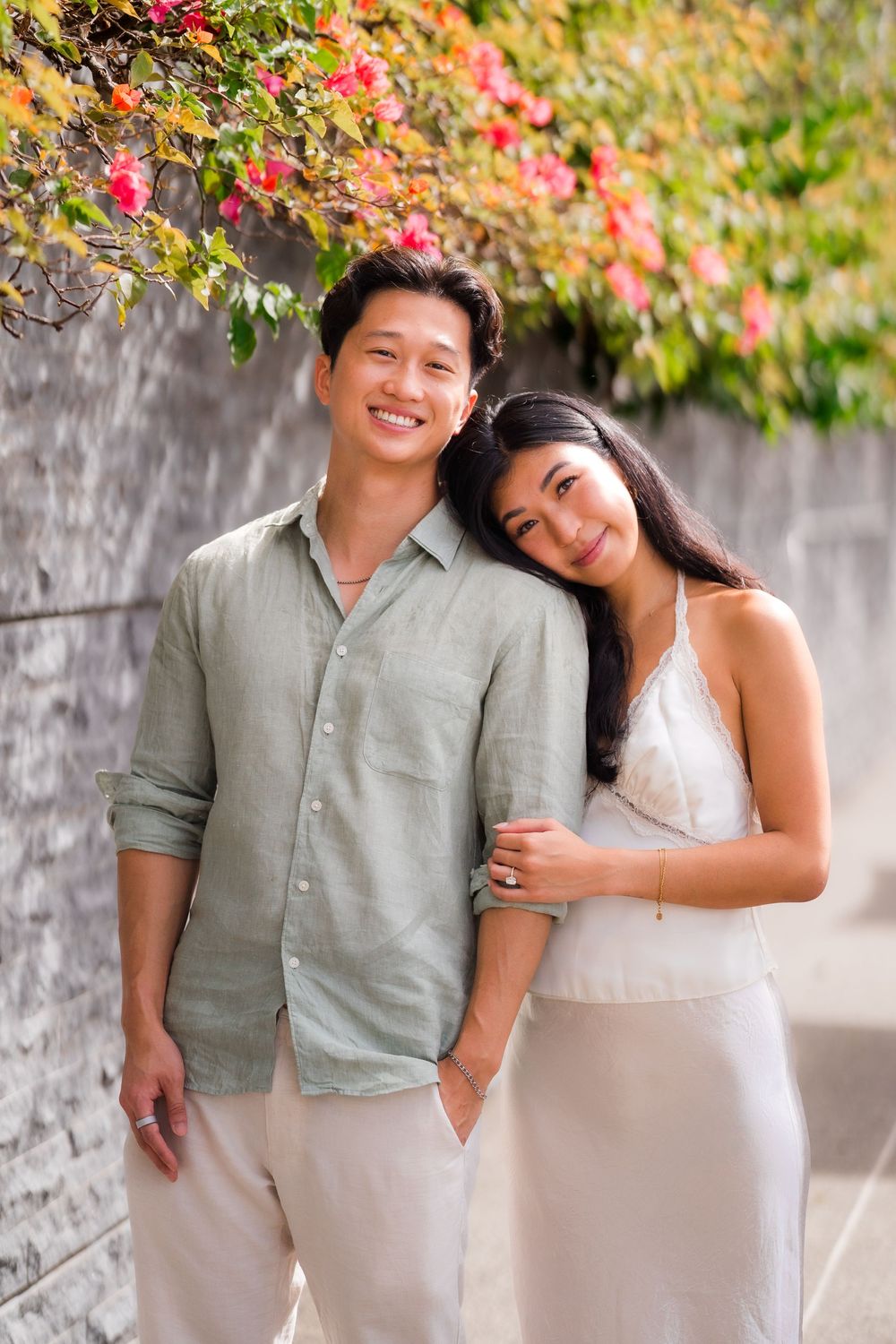 Smiling Asian couple posing together near a stone wall with colorful flowers, man in sage shirt and woman in white dress.