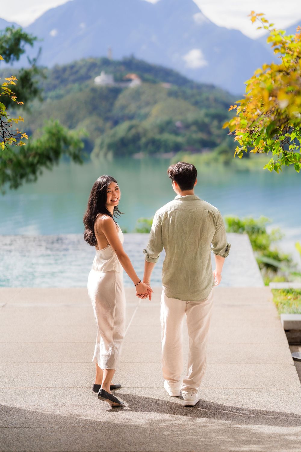 Asian couple holding hands walking toward a scenic mountain lake, woman in white dress looking back and smiling.
