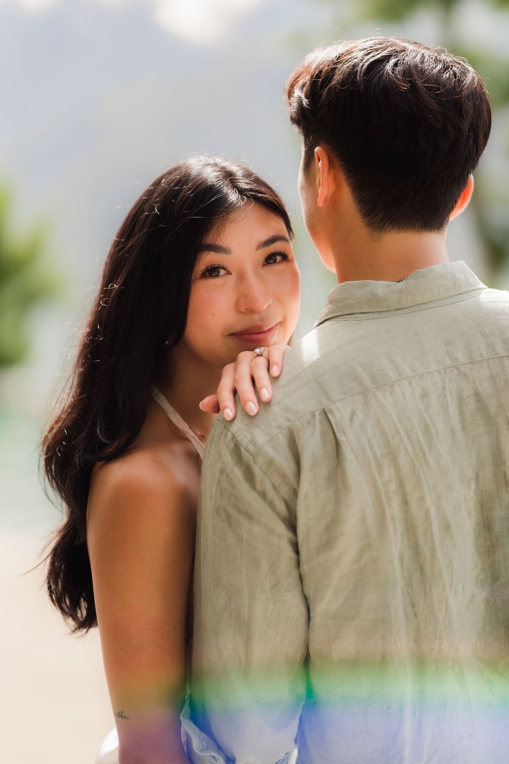 A young Asian couple shares a tender moment outdoors, woman gazing lovingly at man in sage green linen shirt.
