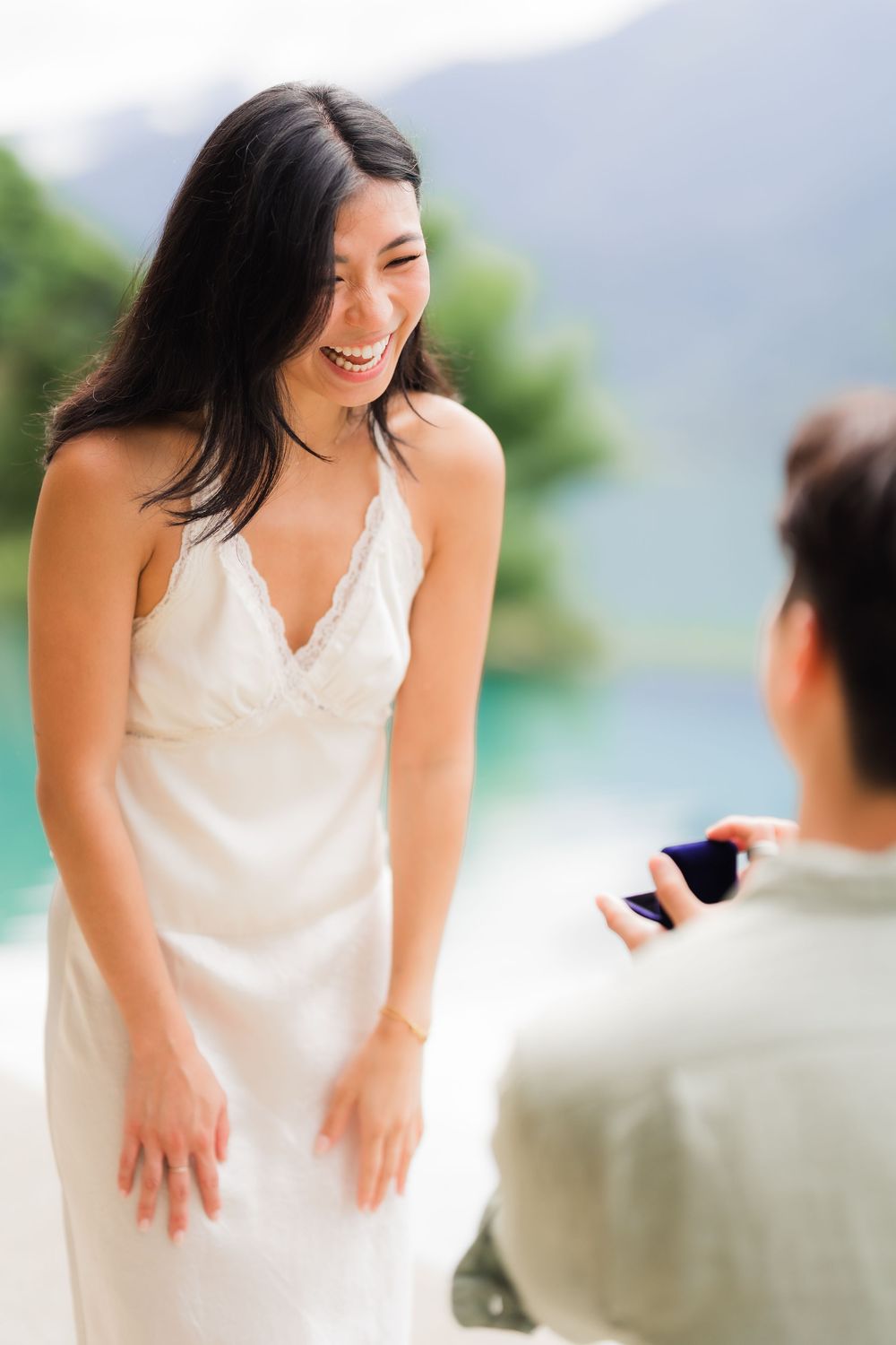 Man proposing to a laughing woman in a white dress on a tropical beach with turquoise water.