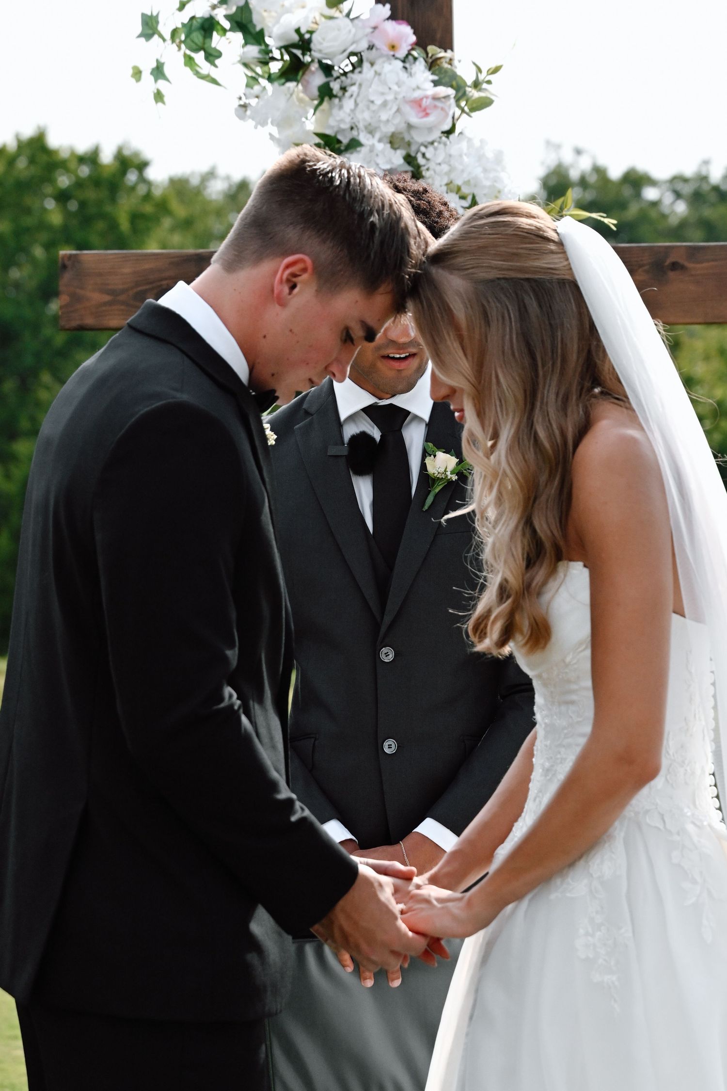 Bride and groom hold hands and bow heads in prayer at outdoor wedding ceremony before floral arch.