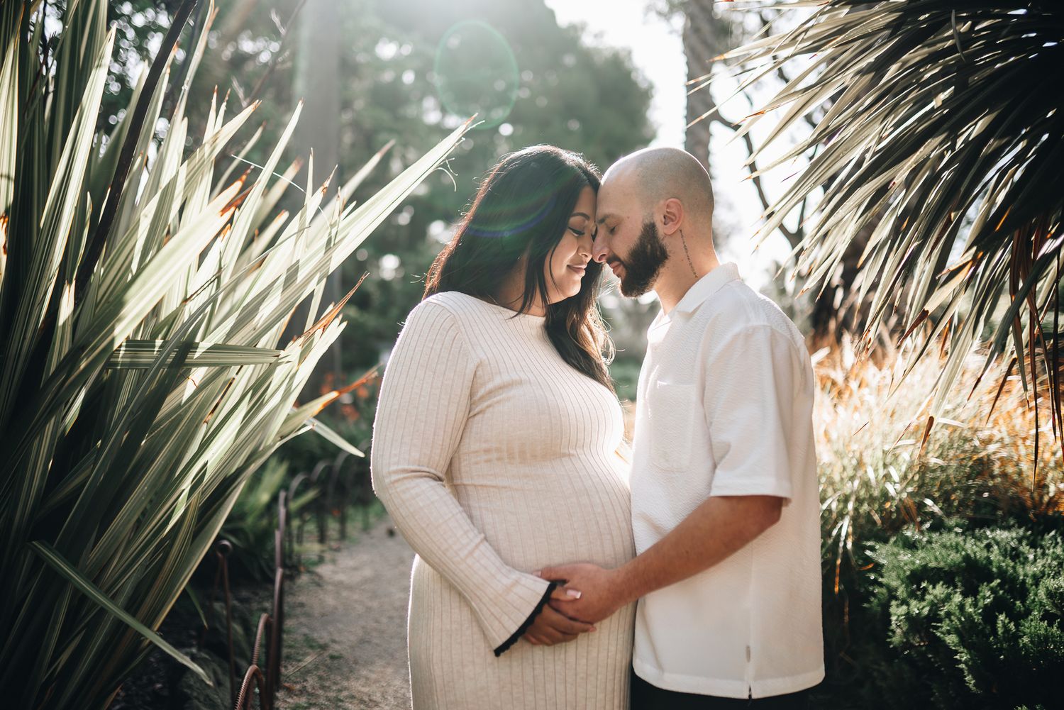 Pregnant couple embracing tenderly in a sunlit garden, surrounded by tropical plants during a Melbourne maternity photo session.