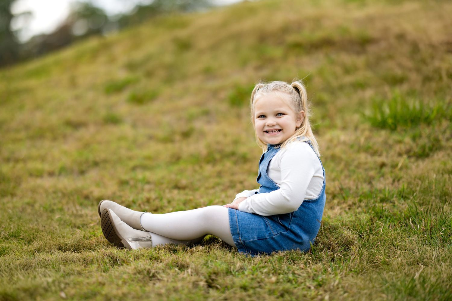 A smiling toddler girl in a denim dress and white tights sits on green grass outdoors.