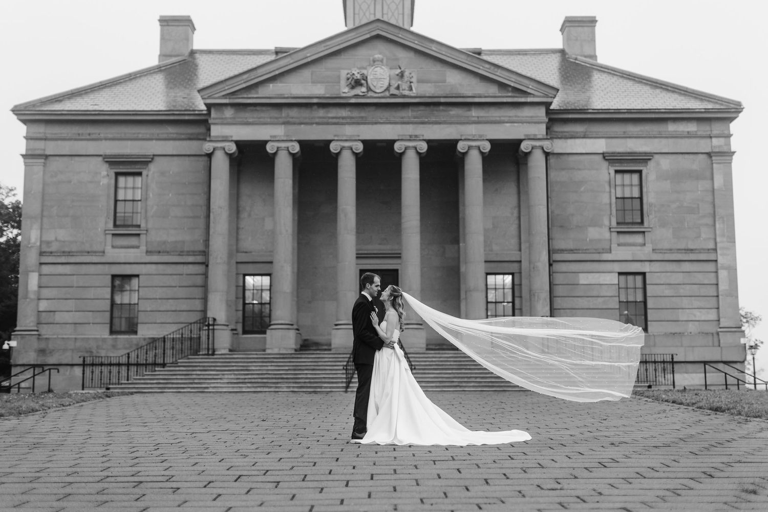 Black and white wedding photo of bride and groom embracing before grand columned building with veil flowing dramatically.