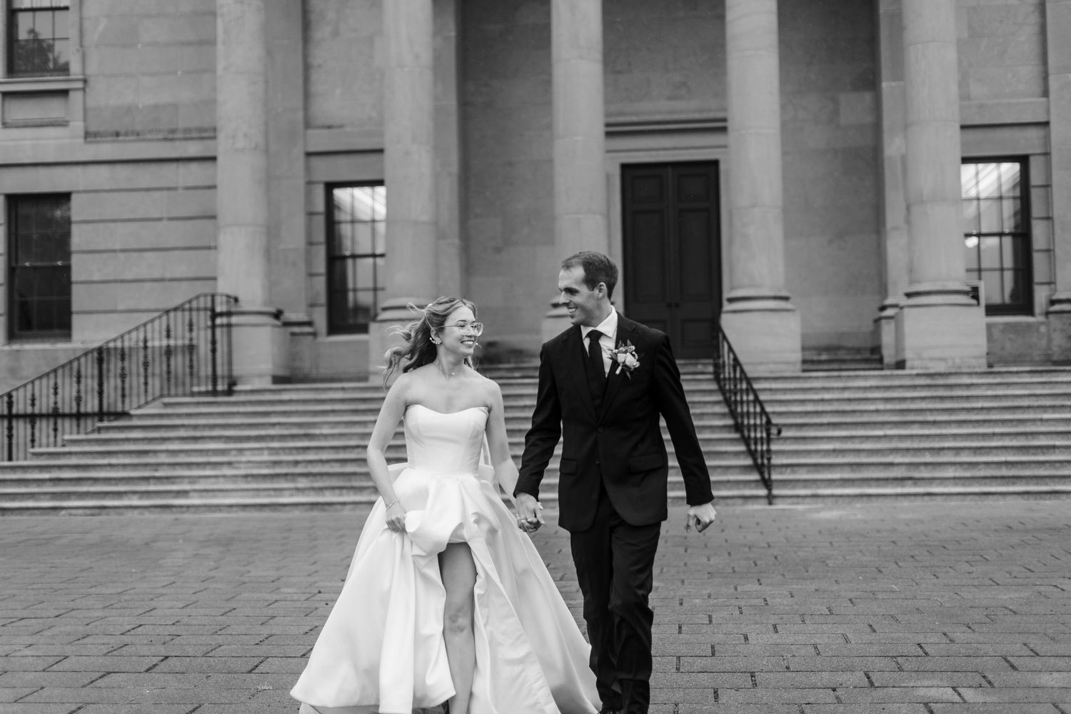 Black and white photo of a bride and groom holding hands, walking and smiling in front of grand columns.