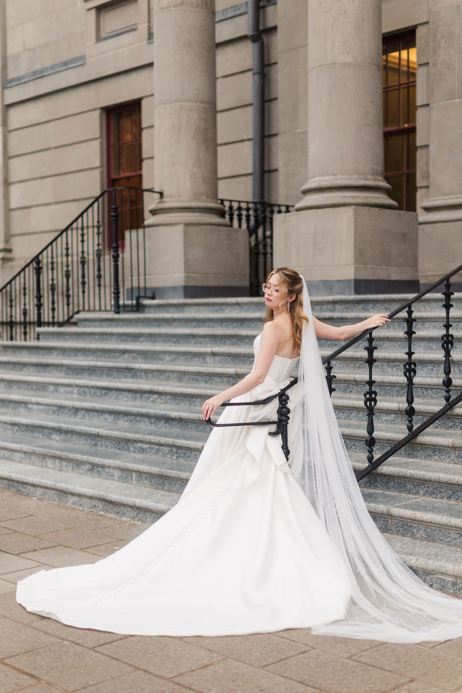 Bride in a white strapless gown with black sash and long veil poses gracefully on stone steps of elegant building.