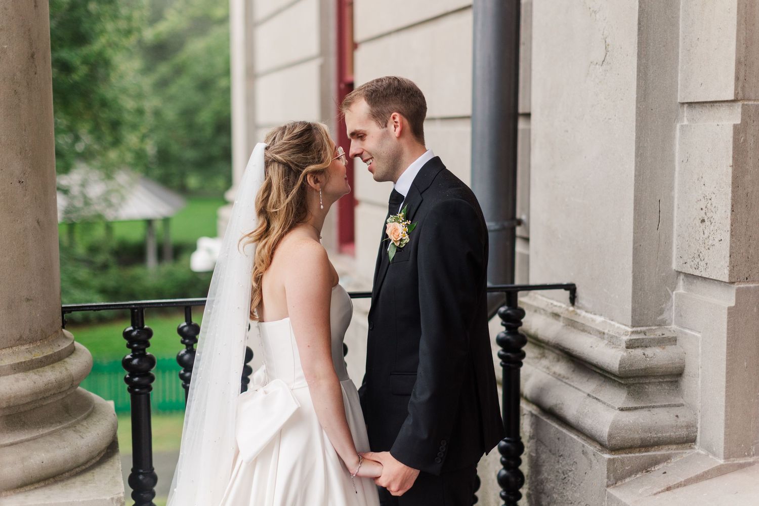 A bride and groom share a romantic moment on a classical stone balcony, holding hands and touching foreheads on their wedding day.
