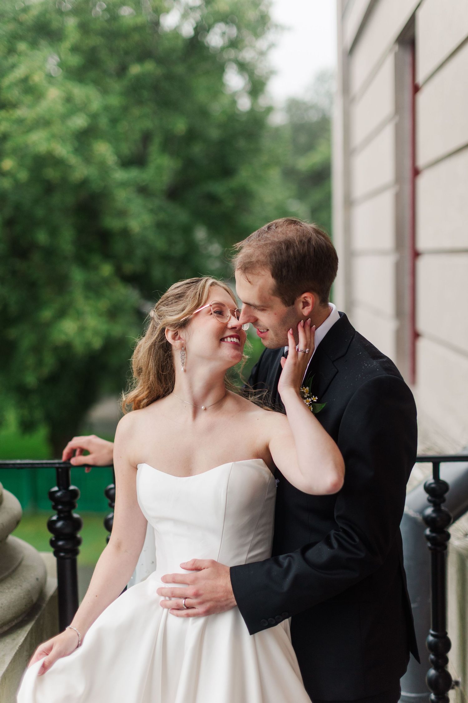 Bride and groom share a romantic moment on a balcony, smiling at each other in wedding attire with green trees behind.